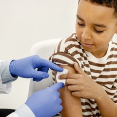 Male doctor holding a cotton on a shoulder. Man wearing white medical gown. Boy wearing brown plaid t-shirt.